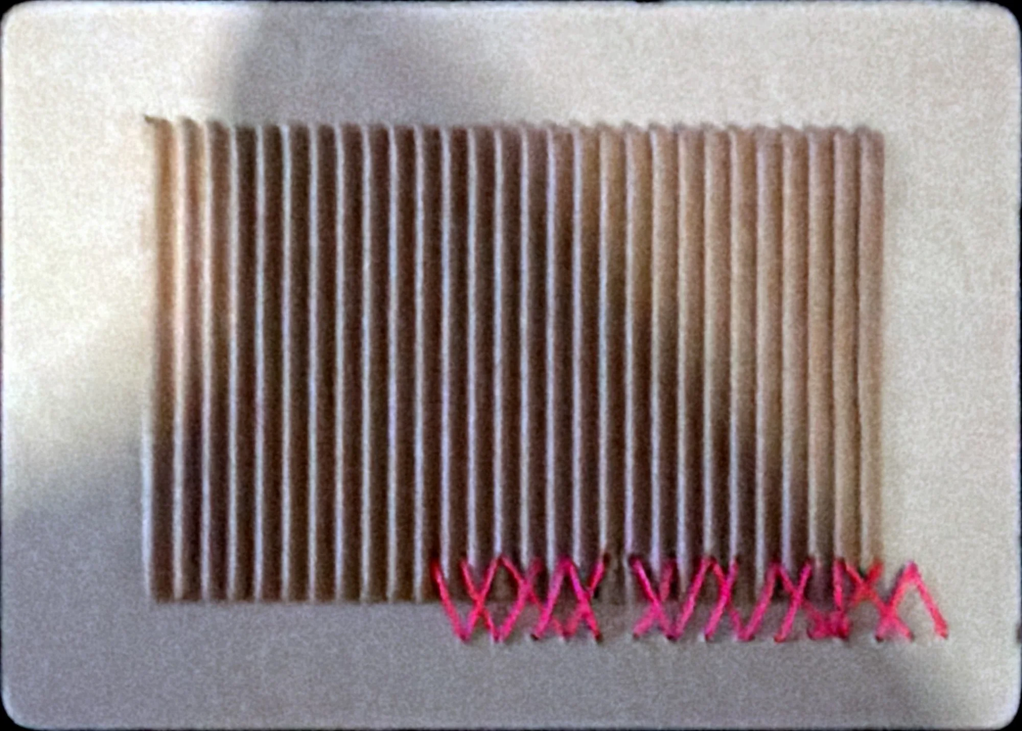 Grey board with rounded corners holding a block of vertical paper pleats, stitched along the bottom with pink cross-stitches.